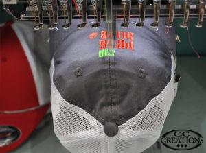 Grey/white cap on an embroidery machine receiving a green and orange embroider logo.